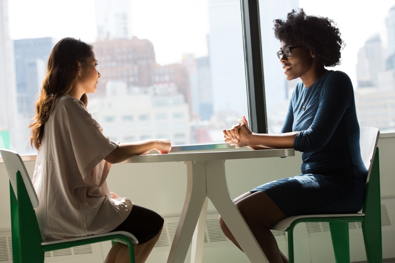 A picture of two women sat either side of a table in formal attire discussing an issue. It looks like an interview